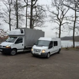 Dwa białe busy transportowe, Renault Master z zabudową kontenerową i Ford Transit, zaparkowane na szutrowym parkingu w pobliżu jeziora i drzew bez liści w pochmurny dzień.