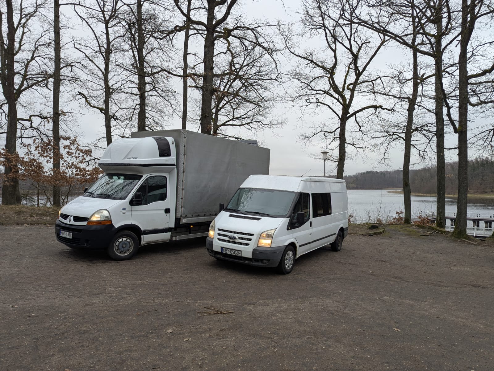 Dwa białe busy transportowe, Renault Master z zabudową kontenerową i Ford Transit, zaparkowane na szutrowym parkingu w pobliżu jeziora i drzew bez liści w pochmurny dzień.