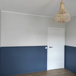 Pokój po remoncie z dwukolorowymi ścianami (szarym i granatowym), drewnianą podłogą, białymi drzwiami z czarną klamką i lampą z wikliny.