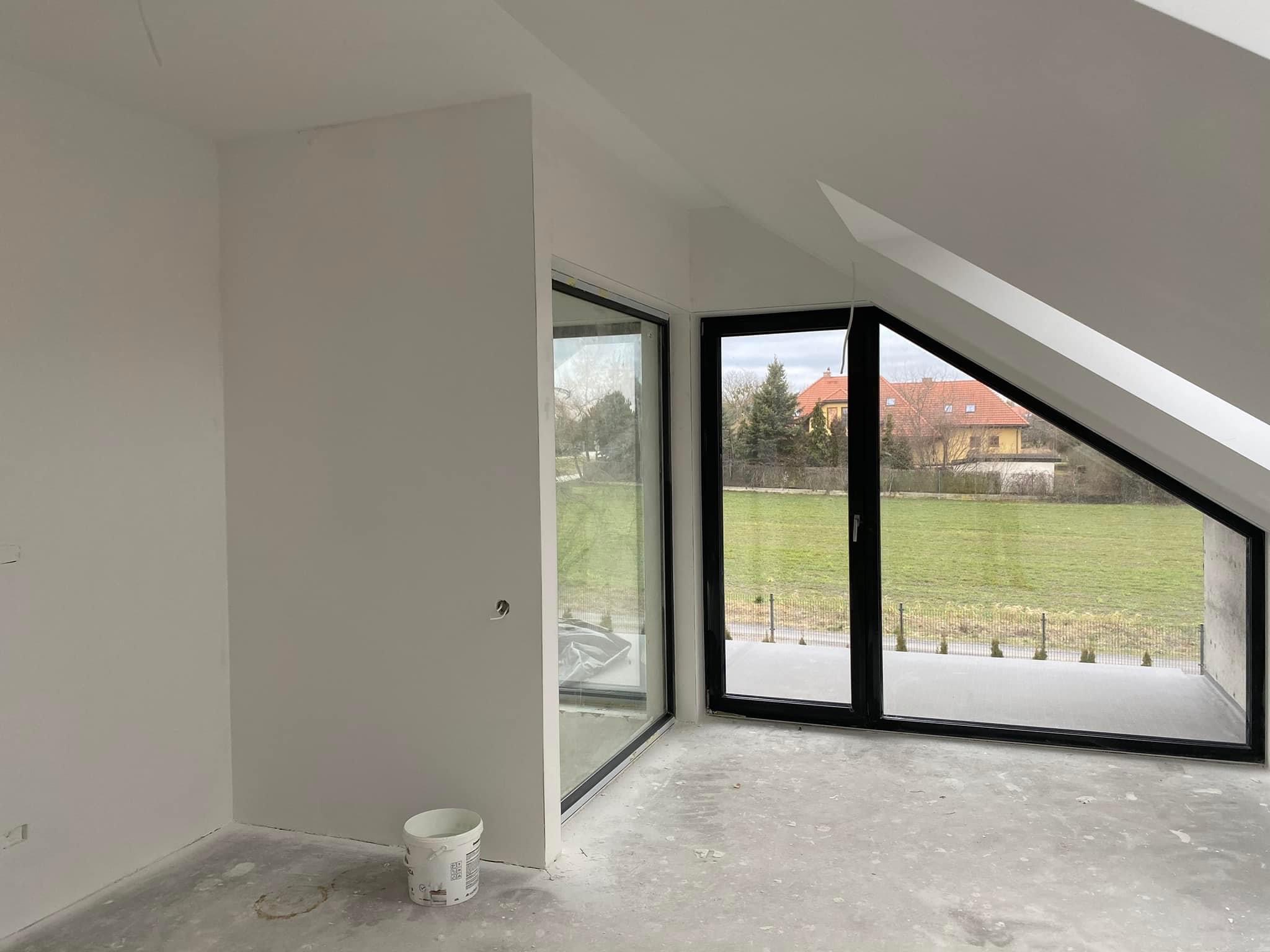 Pokój na poddaszu po gładziowaniu ścian i wylewce, widoczne dwa duże okna z czarnymi ramami i wiaderko z farbą przy ścianie.