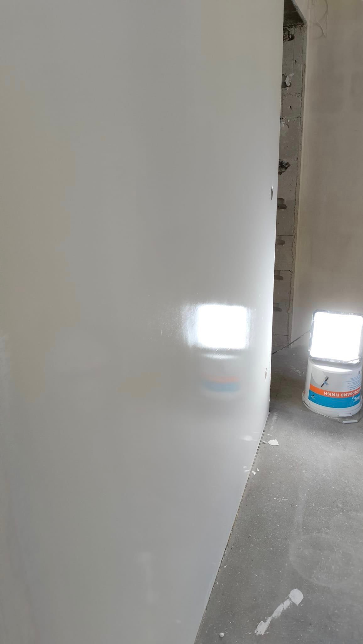 Świeżo pomalowana ściana w jasnym odcieniu szarości, widoczny narożnik i fragment surowej ściany z cegły, na podłodze widoczne krople farby, obok ściany stoi wiadro z farbą i lampa halogenowa.
