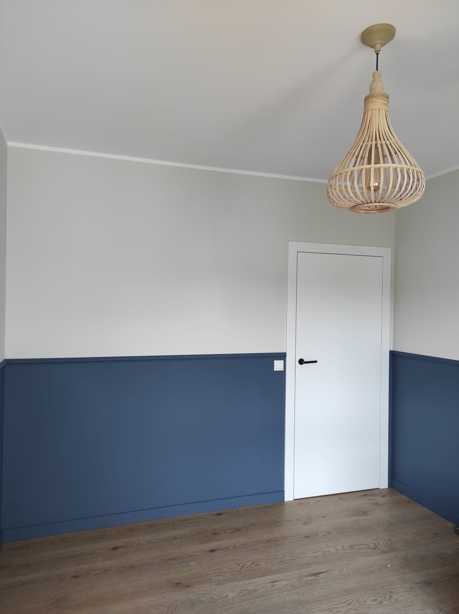 Pokój po remoncie z dwukolorowymi ścianami (szarym i granatowym), drewnianą podłogą, białymi drzwiami z czarną klamką i lampą z wikliny.