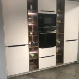 Nowoczesna zabudowa kuchenna z białymi frontami i drewnianymi wnękami, w których znajdują się szklane naczynia, miseczki i wbudowany piekarnik marki Hoover.