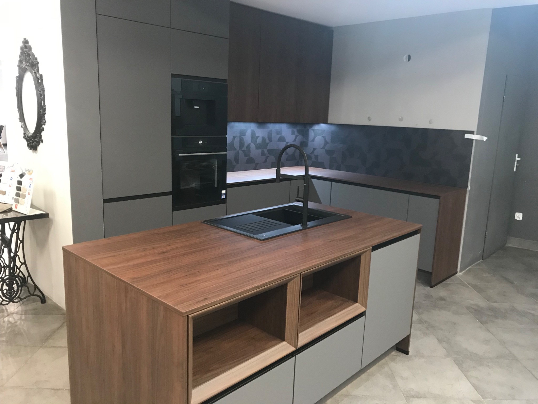 Nowoczesna kuchnia z wyspą, szare szafki z drewnianymi blatami, czarny zlew i bateria, geometryczny backsplash, dekoracyjne lustro na ścianie.