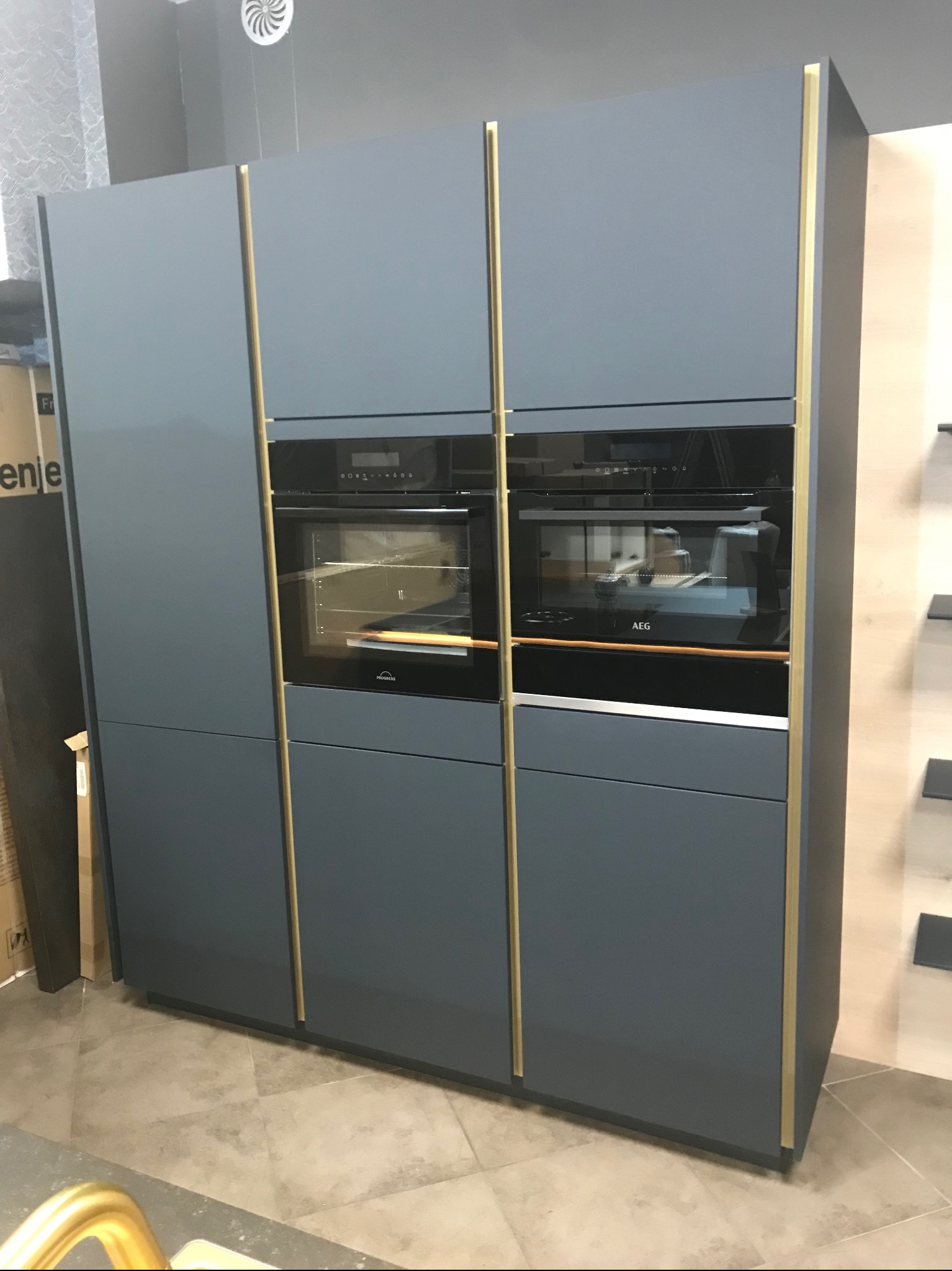 Nowoczesna zabudowa kuchenna w odcieniach szarości z dwoma piekarnikami marki AEG i Progress, wykończona złotymi listwami.