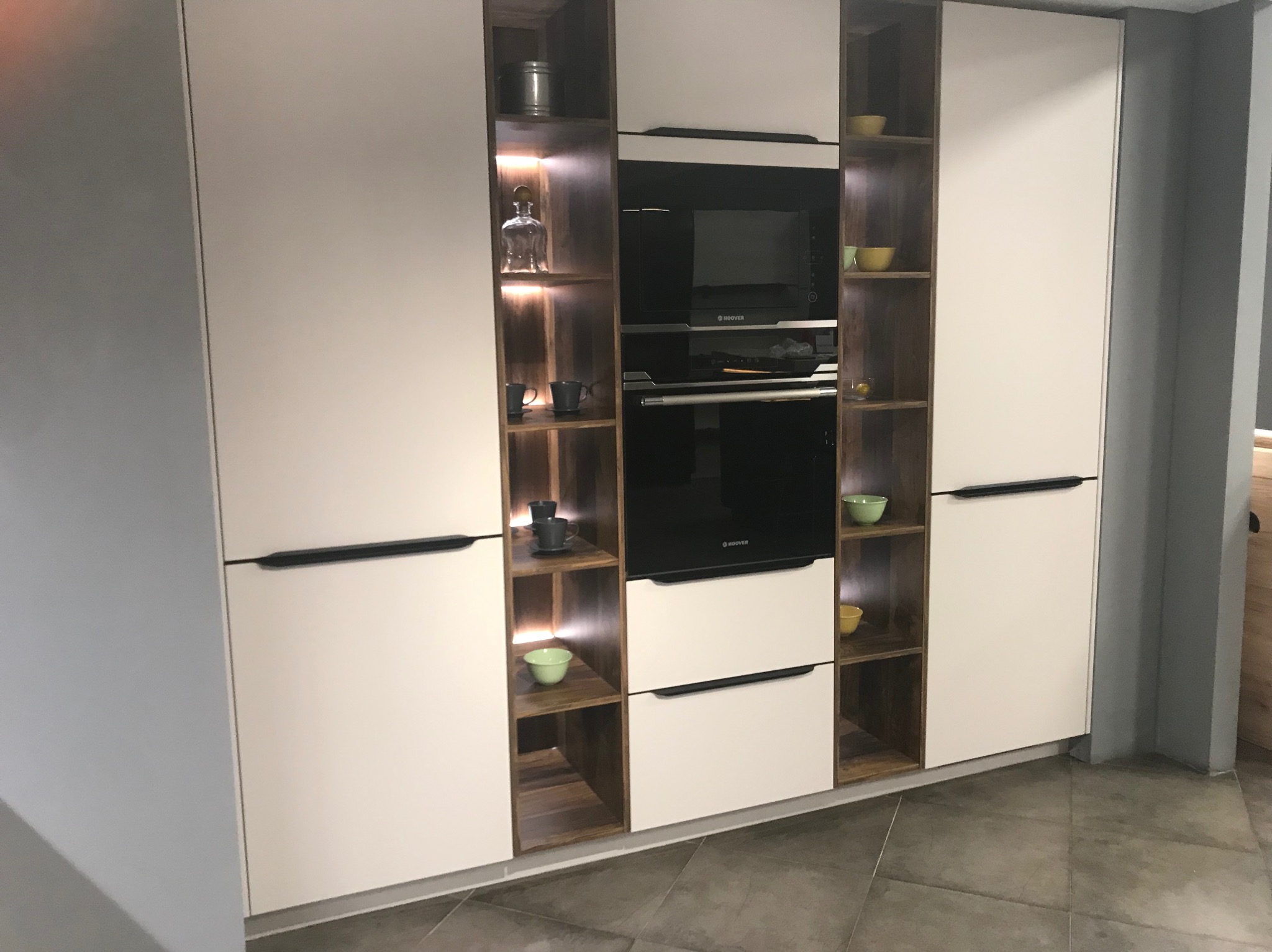 Nowoczesna zabudowa kuchenna z białymi frontami i drewnianymi wnękami, w których znajdują się szklane naczynia, miseczki i wbudowany piekarnik marki Hoover.