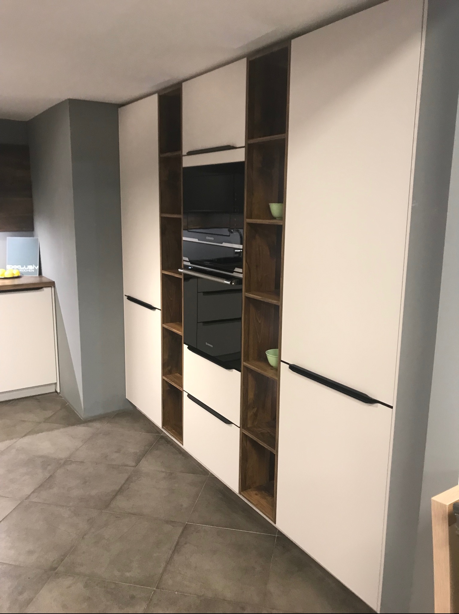 Biała zabudowa kuchenna z ciemnymi drewnianymi wnękami, zawierająca piekarnik i półki z dekoracyjnymi miseczkami, na tle szarej ściany i podłogi z płytek.