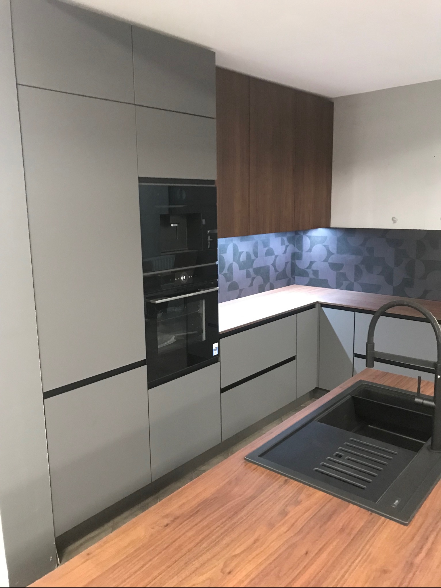 Nowoczesna kuchnia z szarymi, minimalistycznymi szafkami, drewnianym blatem i czarnym zlewem z baterią; podświetlenie LED nad blatem roboczym.