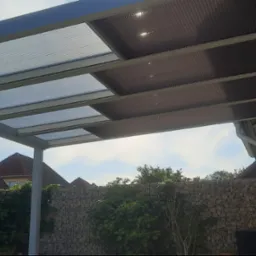 Metalowa konstrukcja z przezroczystym zadaszeniem, widoczna z dołu, na tle zieleni i kamiennego muru.