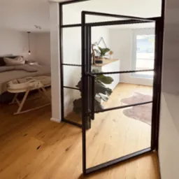 Nowoczesne, przeszklone drzwi loftowe z czarnymi szprosami oddzielają sypialnię z kołyską od jasnego wnętrza z drewnianą podłogą i elementami dekoracyjnymi.