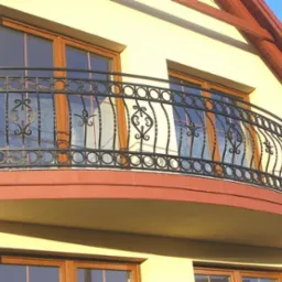 Elegancki balkon z owalną, kutą, czarną balustradą z dekoracyjnymi elementami na tle jasnej elewacji domu z drewnianymi oknami i widocznym fragmentem dachu.