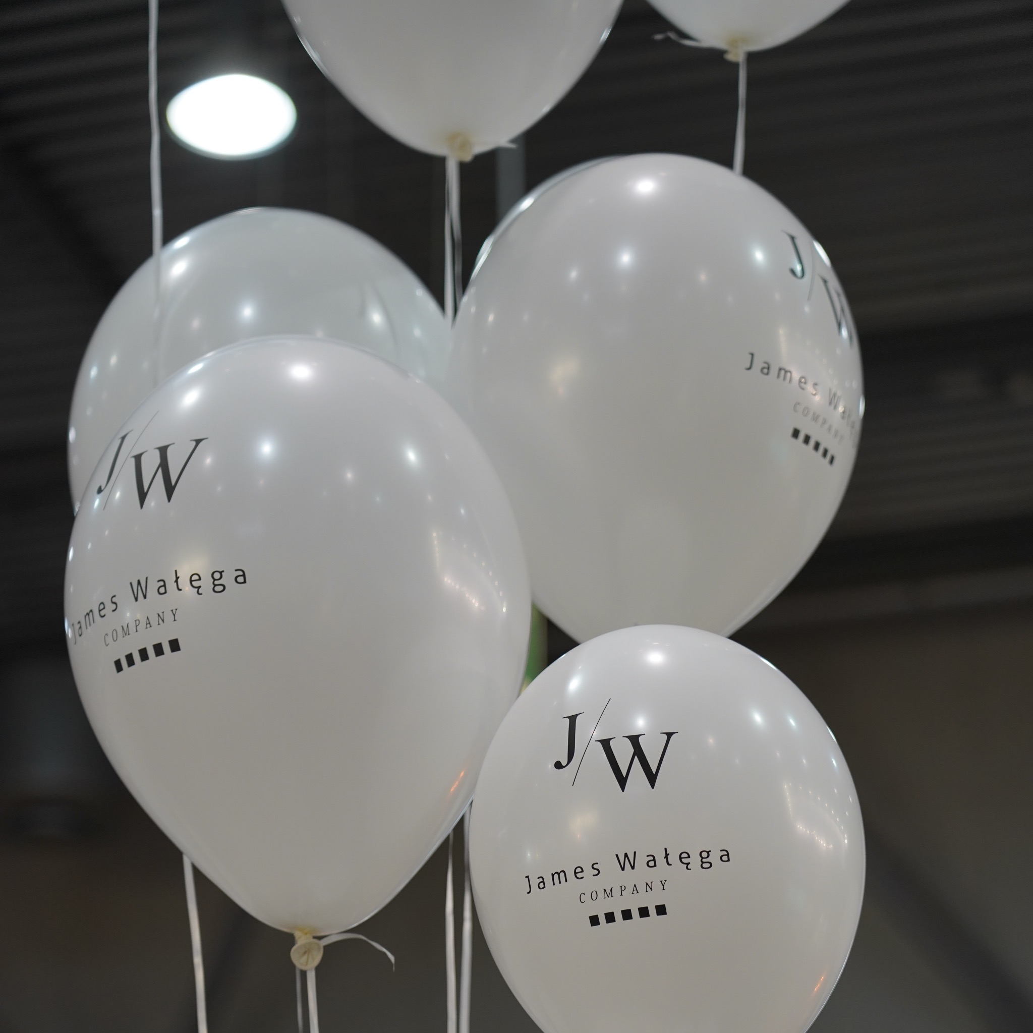 Białe balony z nadrukowanym logo firmy James Wałęga Company, wiszące na przezroczystych żyłkach, oświetlone od góry.