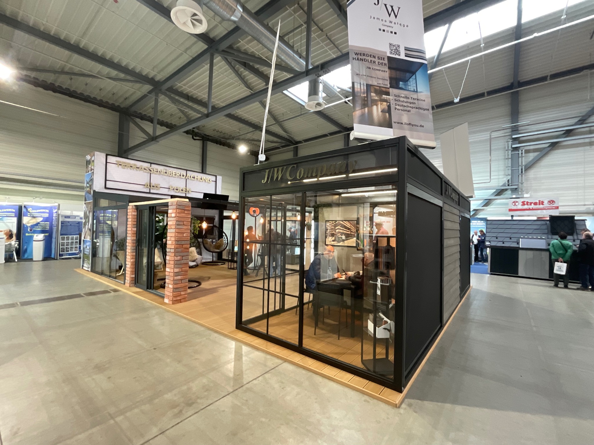 Ekspozycja stoiska targowego z przeszkloną konstrukcją z czarnymi ramami, prezentująca firmę J/W Company z wiszącym banerem reklamowym i elementami wykończenia z cegły.