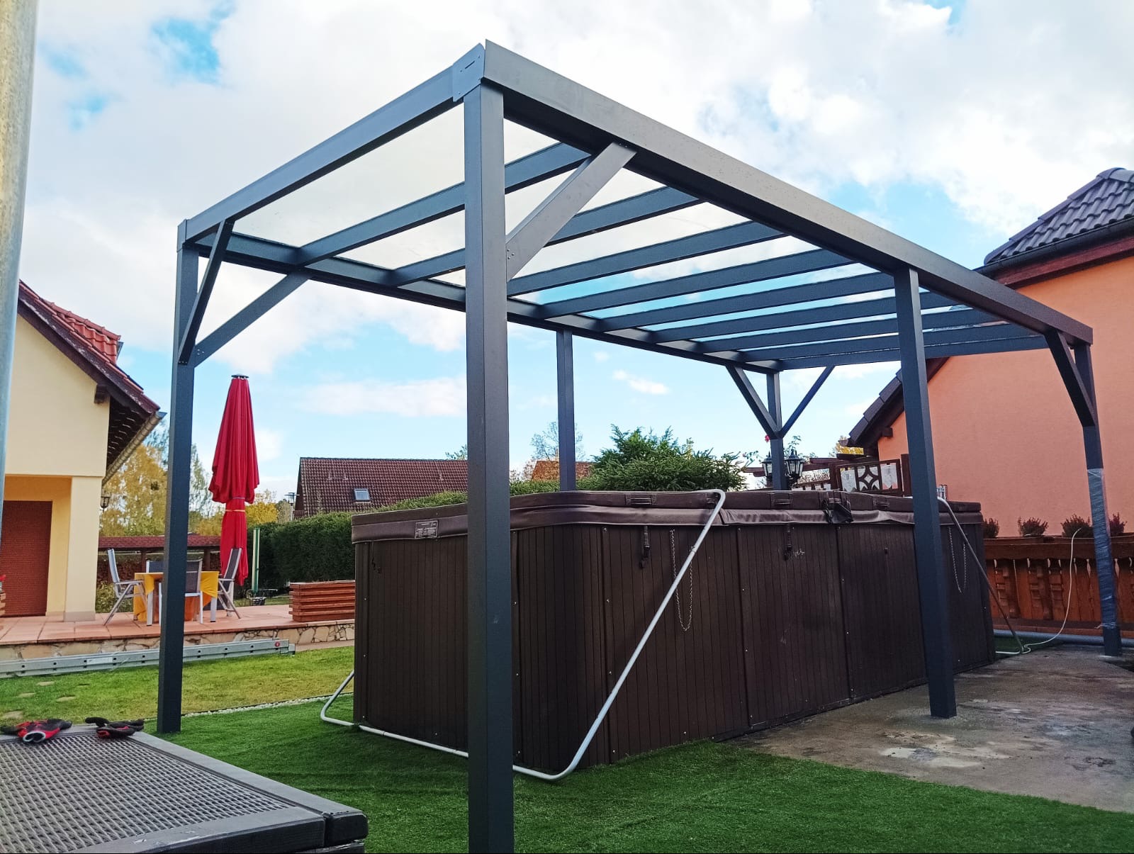 Nowoczesna, grafitowa konstrukcja z przezroczystym zadaszeniem, osłaniająca brązowe jacuzzi na sztucznej trawie w ogrodzie, z widocznym czerwonym parasolem i elementami zabudowy w tle.