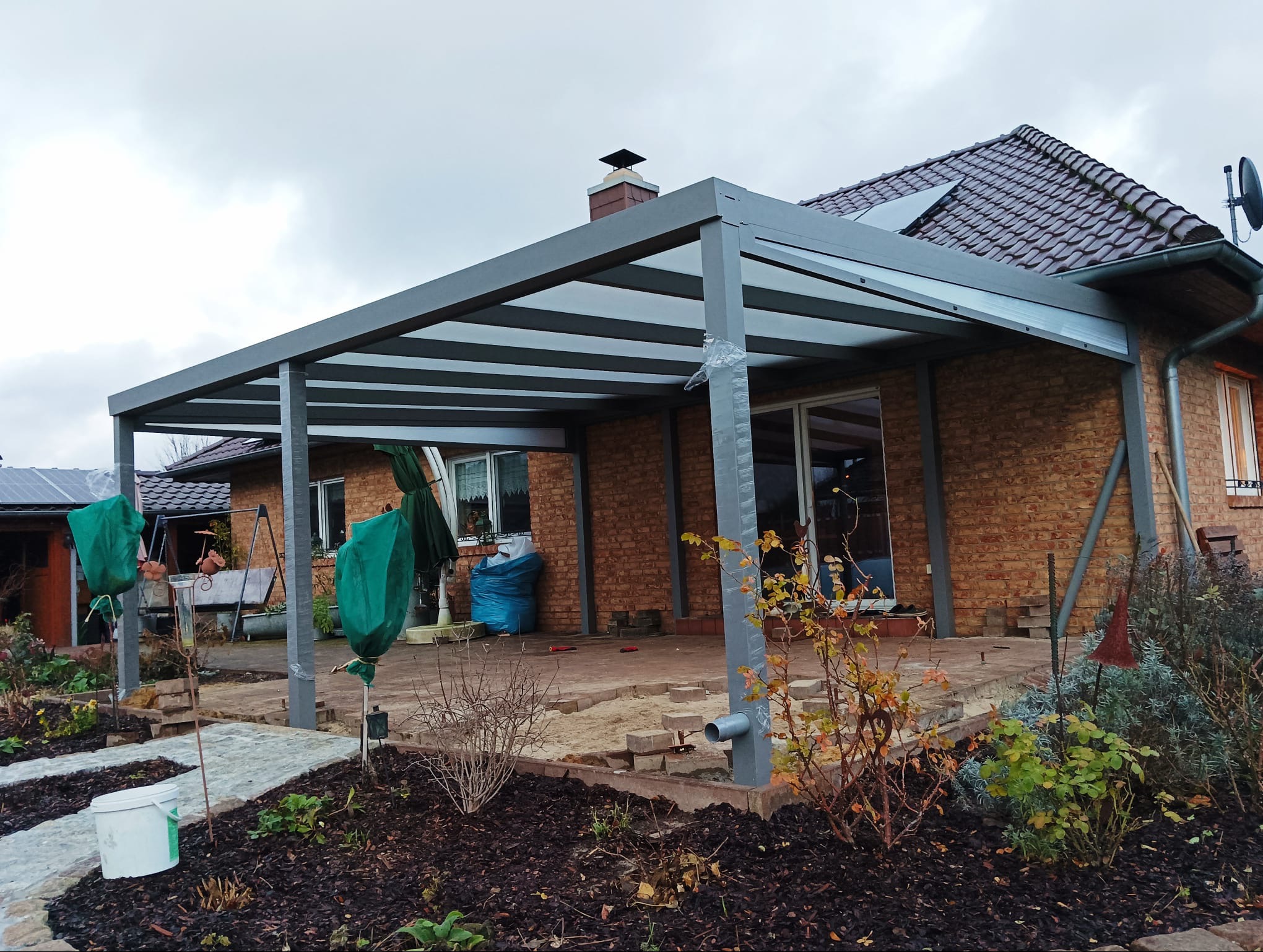 Szara pergola metalowa z przezroczystym zadaszeniem przy ceglanym domu, widoczne prace brukarskie na tarasie i zabezpieczone rośliny w ogrodzie.
