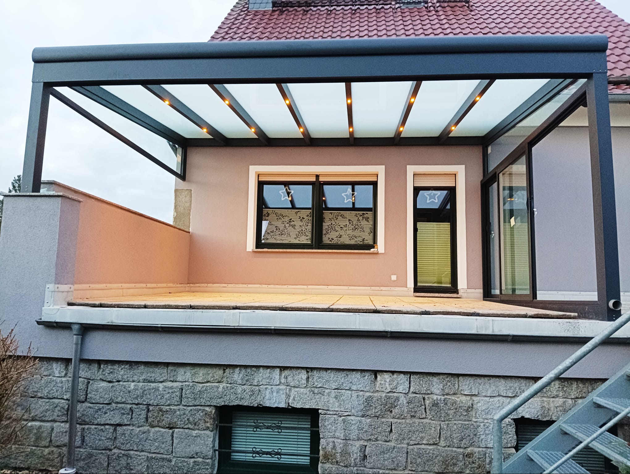 Nowoczesna pergola metalowa z przeszklonym dachem i punktowym oświetleniem LED, przylegająca do budynku z oknami ozdobionymi gwiazdkami.