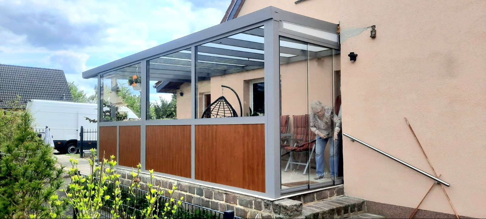 Zadaszony taras z przesuwnymi szklanymi drzwiami i drewnianymi panelami, widoczna osoba wewnątrz, wiszące krzesło i kwiaty w doniczkach.