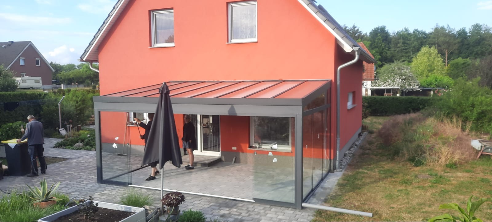 Szklana zabudowa tarasu z metalową konstrukcją, przylegająca do czerwonego domu jednorodzinnego, widoczni ludzie wewnątrz i na zewnątrz konstrukcji.