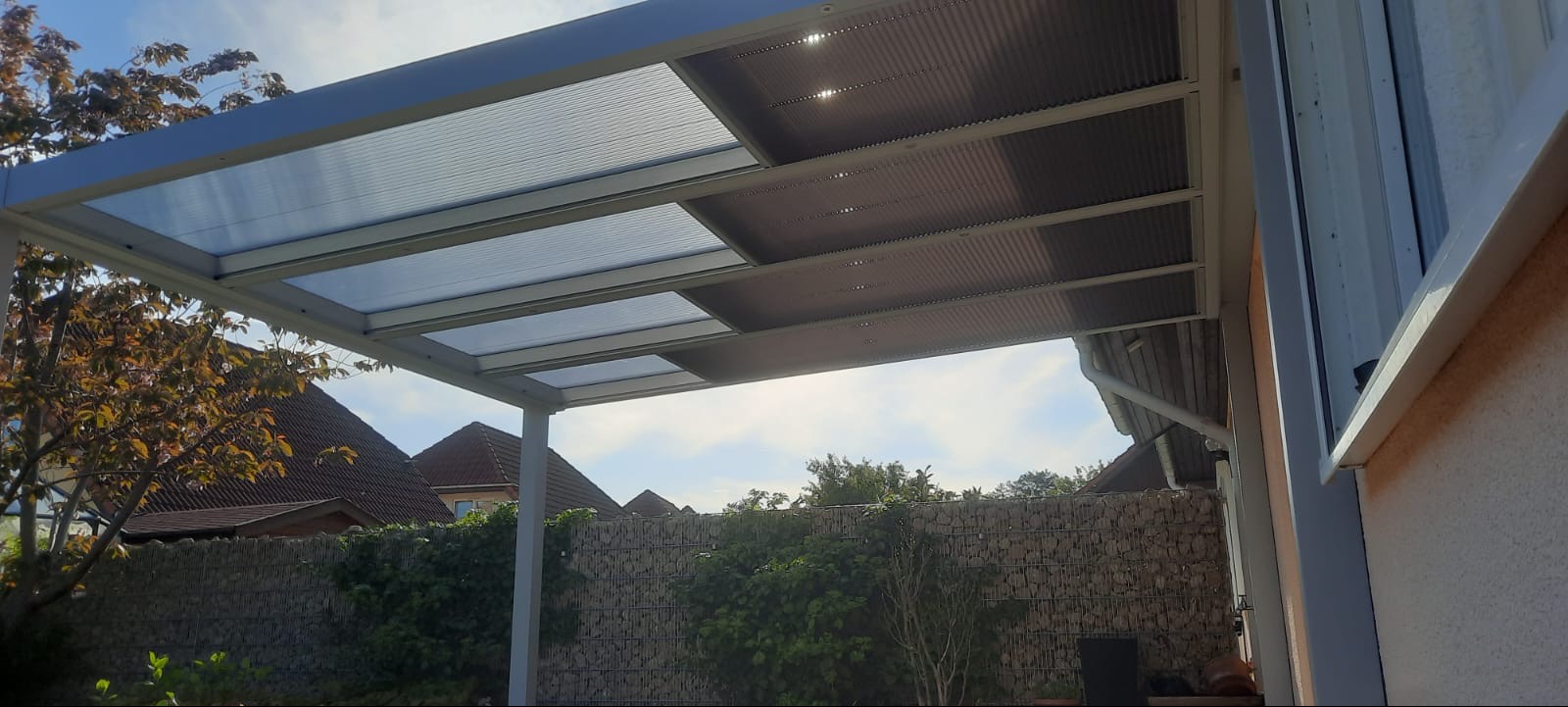 Metalowa konstrukcja z przezroczystym zadaszeniem, widoczna z dołu, na tle zieleni i kamiennego muru.