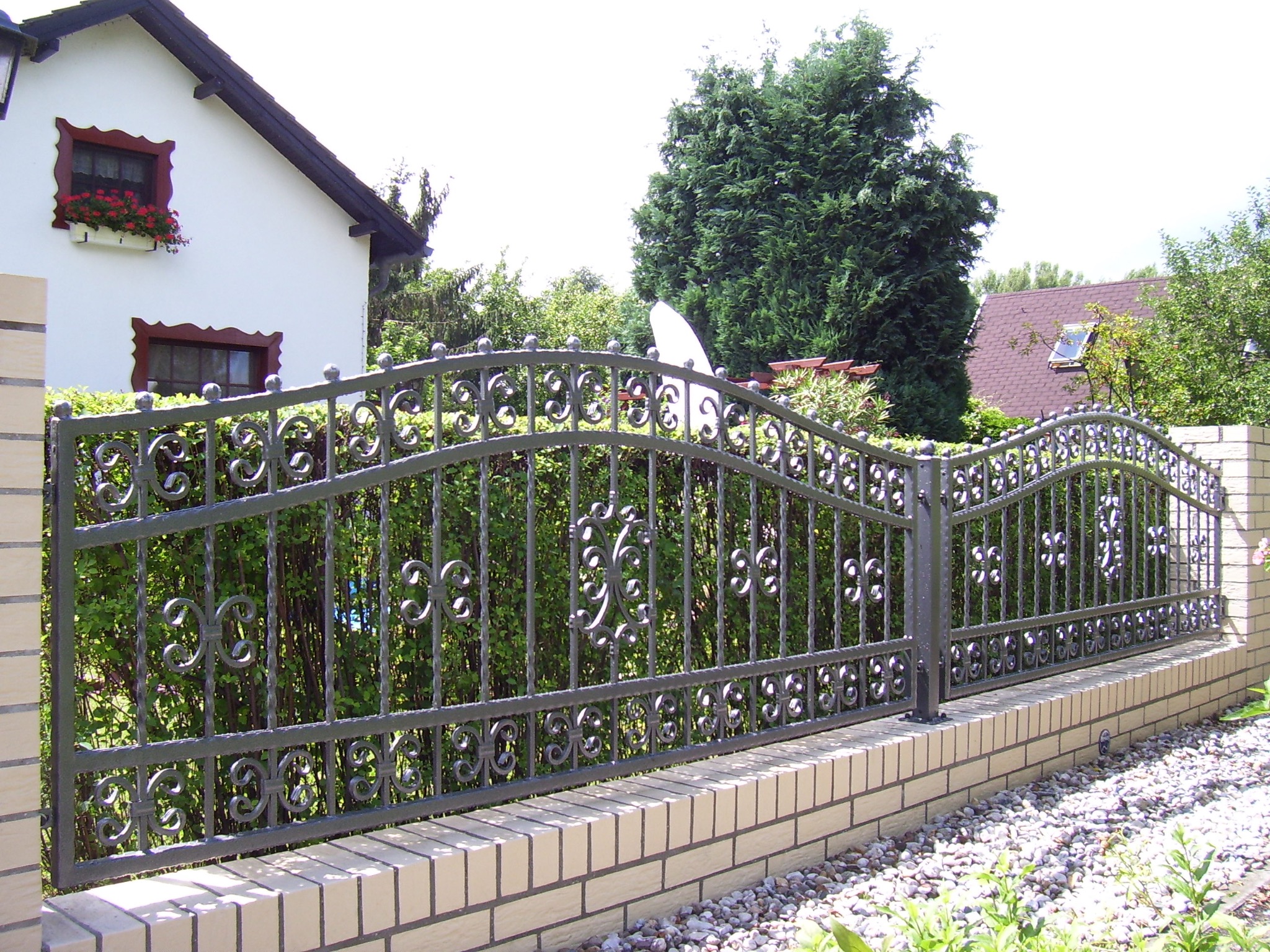 Kuta, metalowa, segmentowa brama wjazdowa z dekoracyjnymi elementami, osadzona na niskim, ceglanym murku, na tle domu z oknami zdobionymi kwiatami i zielenią.