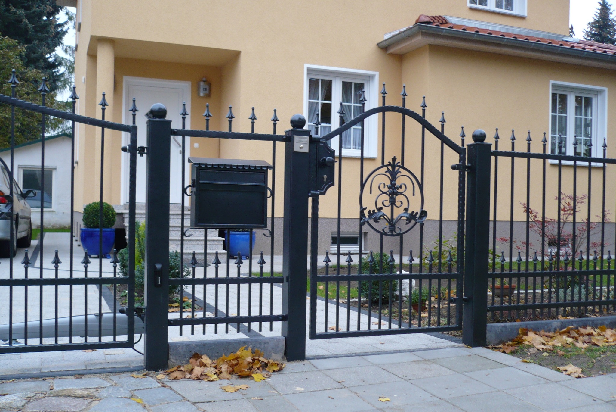 Czarna, metalowa brama wjazdowa z dekoracyjnym ornamentem i skrzynką na listy, prowadząca do domu z żółtą elewacją i białymi oknami.