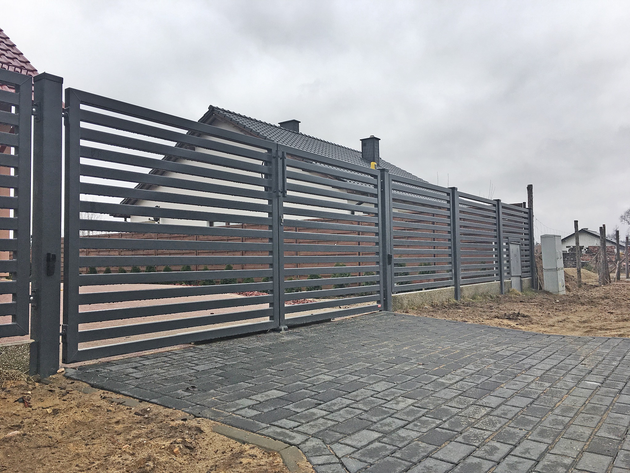 Nowoczesne, poziome ogrodzenie z metalowych paneli w kolorze grafitowym, z bramą wjazdową, na posesji z kostką brukową i domem w tle, pochmurny dzień.