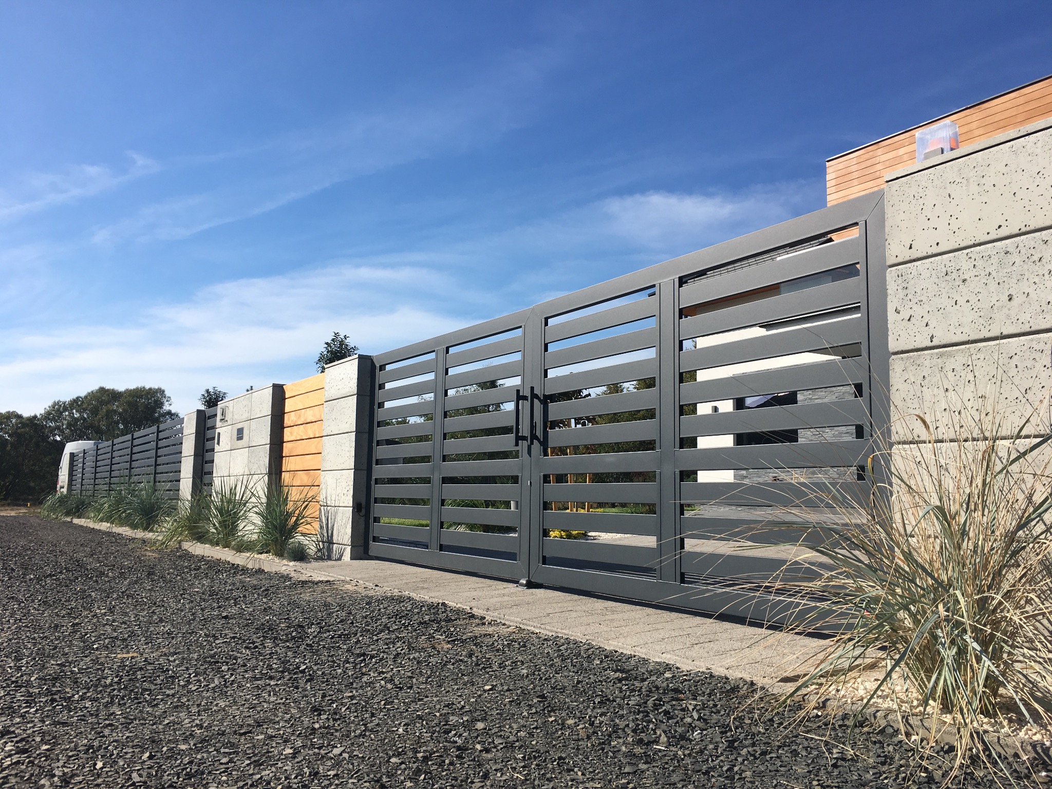 Nowoczesna, pozioma brama wjazdowa z szarymi, metalowymi przęsłami, osadzona w betonowych słupkach, widok z zewnątrz na tle błękitnego nieba.