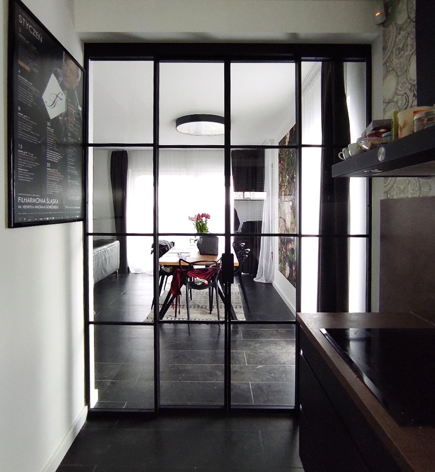 Nowoczesne, czarne, stalowe drzwi wewnętrzne z licznymi szybami oddzielające kuchnię od jadalni, widok na stół z krzesłami i salon z sofą, plakat na ścianie po lewej stronie.