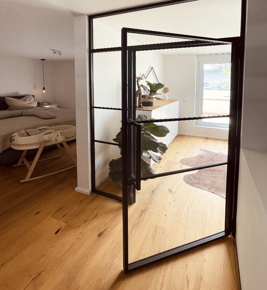 Nowoczesne, przeszklone drzwi loftowe z czarnymi szprosami oddzielają sypialnię z kołyską od jasnego wnętrza z drewnianą podłogą i elementami dekoracyjnymi.