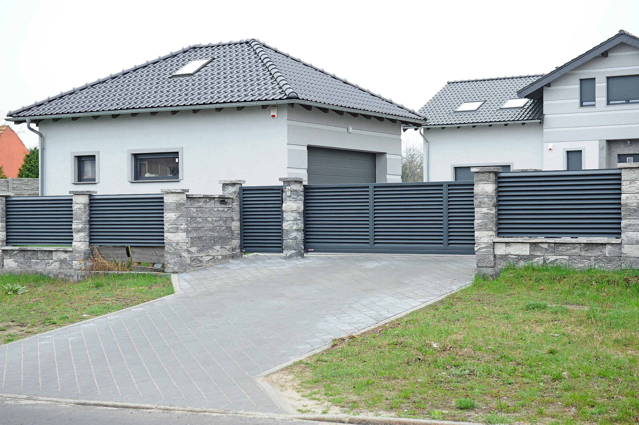 Nowoczesna posesja z przesuwną bramą i ogrodzeniem panelowym w kolorze grafitowym, wykończonym kamiennymi słupkami, prowadząca do domu z szarą dachówką i oknami z roletami zewnętrznymi.