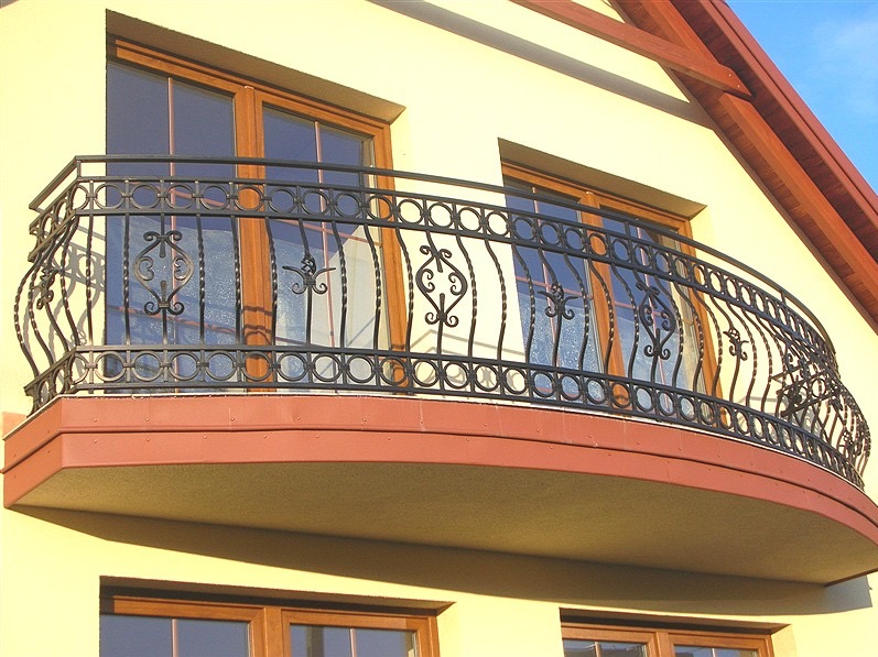 Elegancki balkon z owalną, kutą, czarną balustradą z dekoracyjnymi elementami na tle jasnej elewacji domu z drewnianymi oknami i widocznym fragmentem dachu.