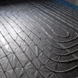 Białe rury ułożone w pętle na srebrnej folii z czerwoną siatką, tworzące system ogrzewania podłogowego w pomieszczeniu w trakcie instalacji.