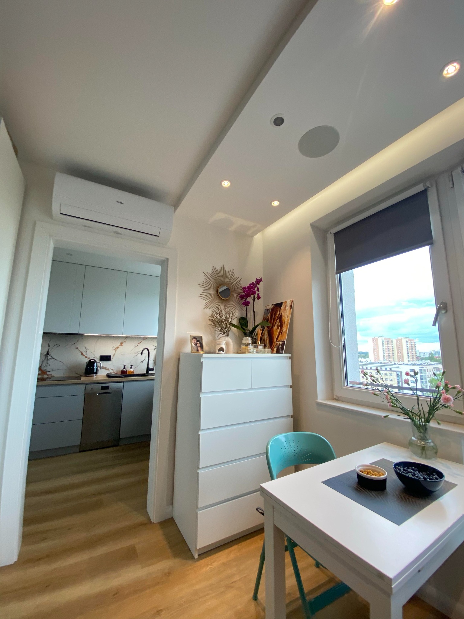 Nowoczesne wnętrze mieszkania w Koronowie z klimatyzacją, widok na kuchnię z drewnianą podłogą, białą komodę z dekoracjami i stolik z owocami przy oknie.