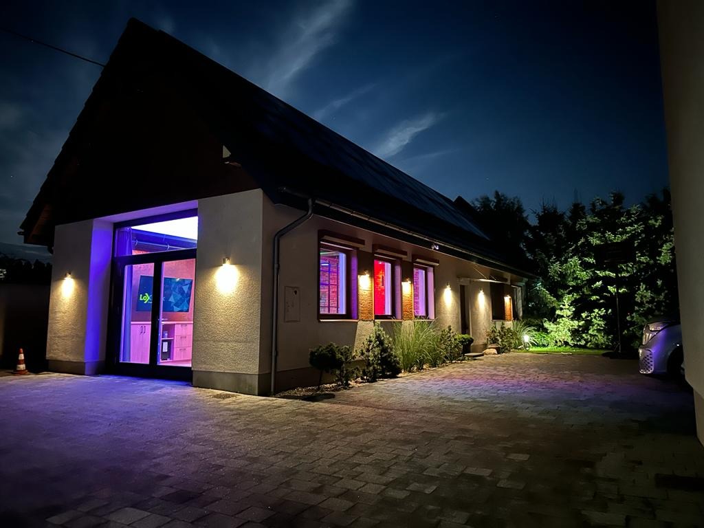 Nocny widok domu jednorodzinnego z oświetleniem LED w kolorze fioletowym i czerwonym, oświetlającym okna i wejście. Widoczny podjazd z kostki brukowej i fragment samochodu.