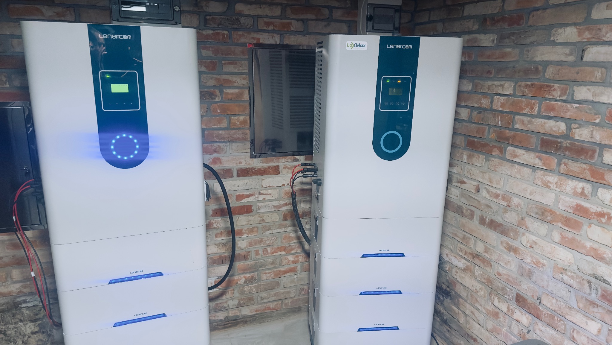 Dwa białe magazyny energii Lenercom z niebieskimi akcentami, zainstalowane w pomieszczeniu z ceglaną ścianą w Koronowie. Widoczne przewody i wyświetlacze LED na urządzeniach.