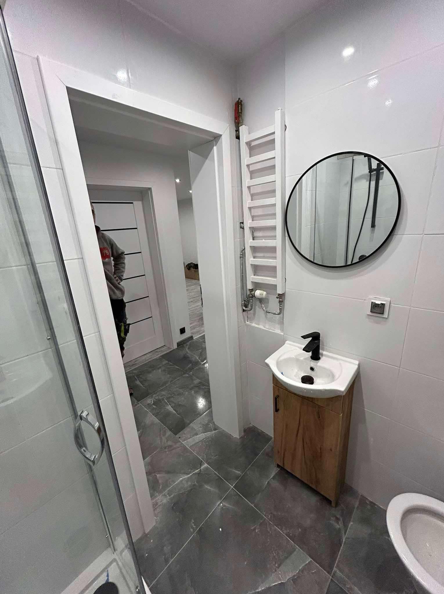 Nowoczesna łazienka z szarymi płytkami podłogowymi, białe płytki ścienne, umywalka z szafką z drewnopodobnym wykończeniem i okrągłe lustro w czarnej ramie, widoczny grzejnik drabinkowy.