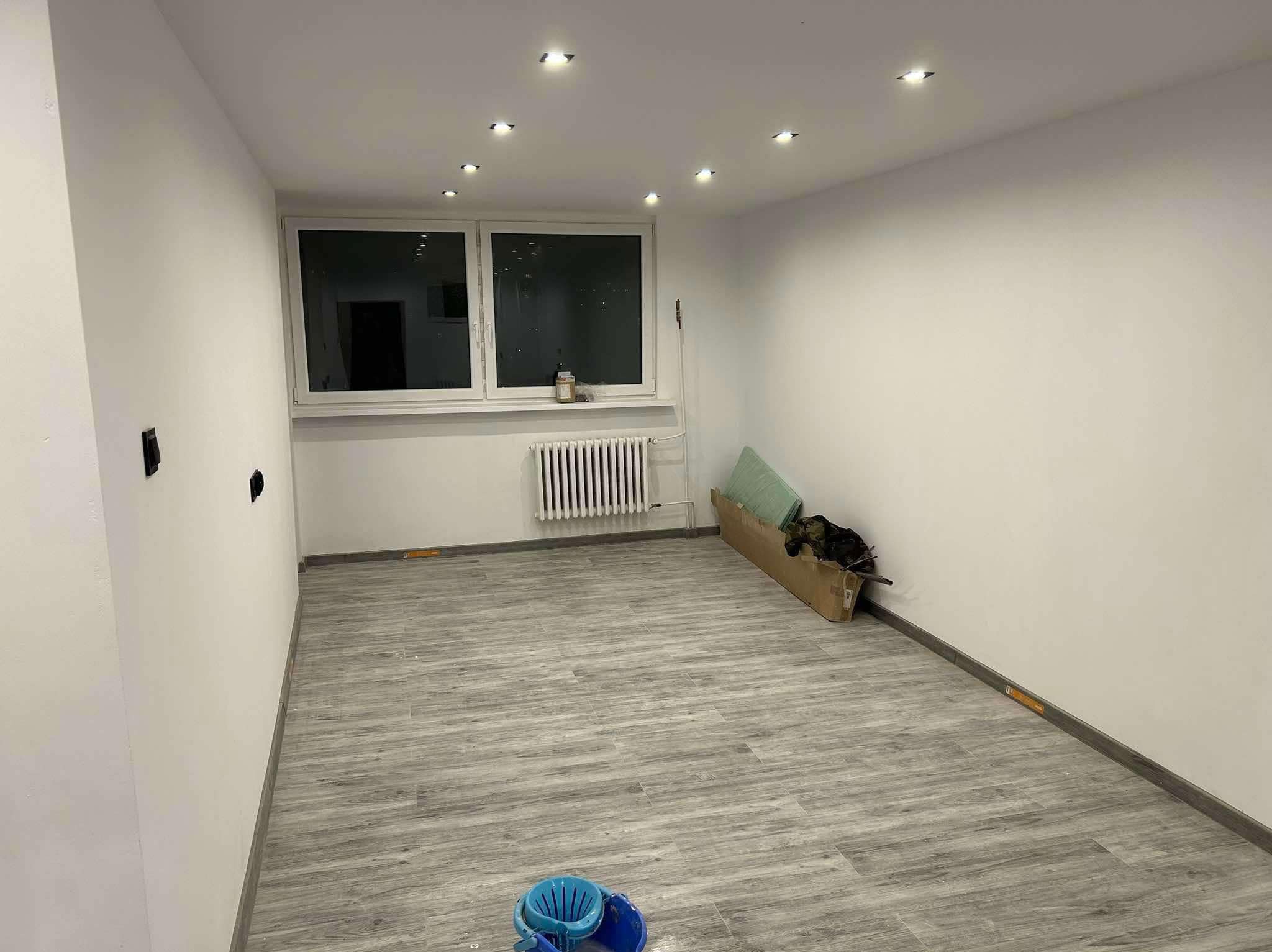 Pusty pokój po odświeżeniu: świeżo pomalowane białe ściany, szare panele podłogowe imitujące drewno, sufit z punktowym oświetleniem LED, grzejnik pod oknem, karton z materiałami budowlanymi przy...