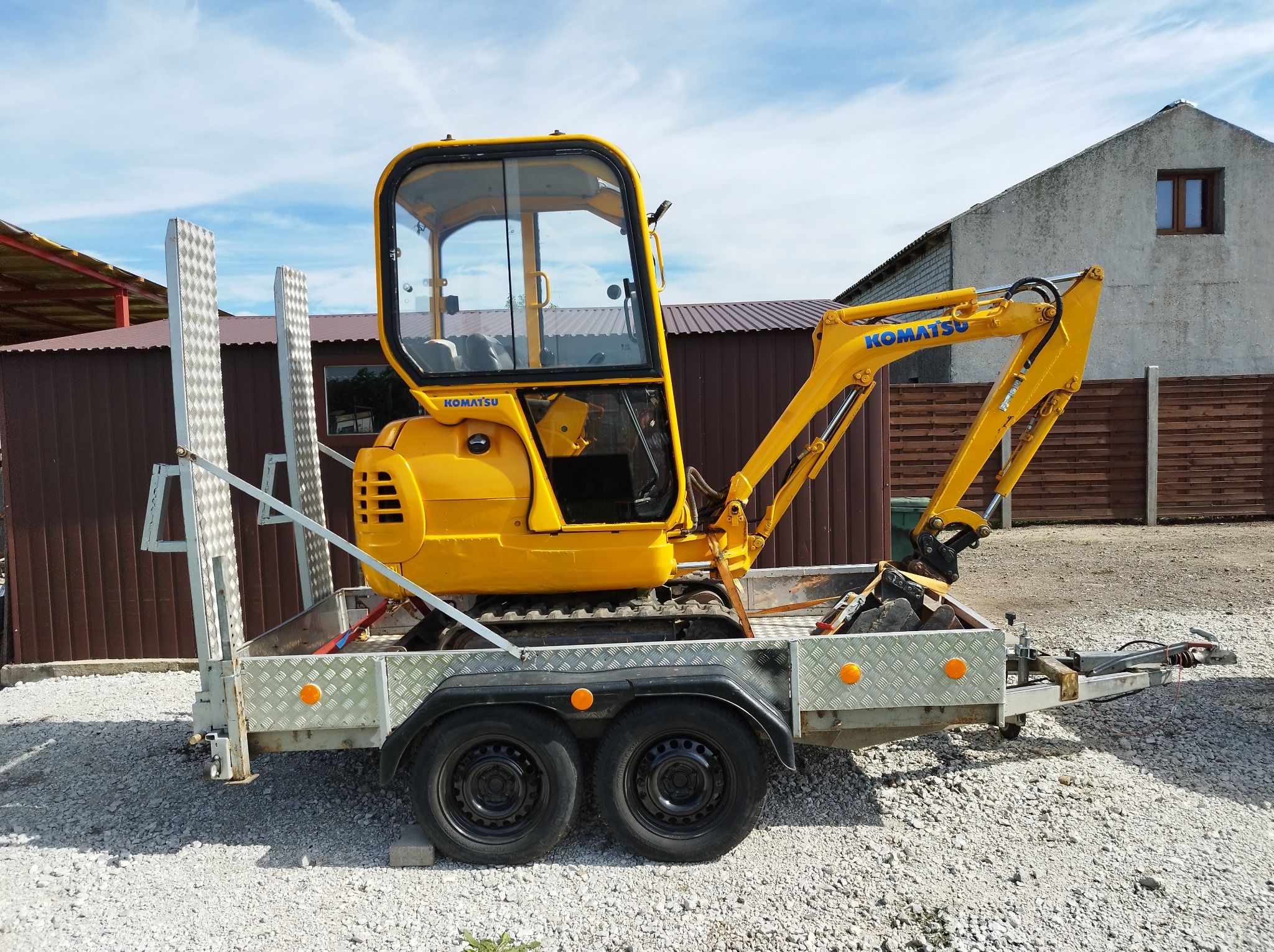 Żółta minikoparka Komatsu na przyczepie z najazdami, zaparkowana na żwirowym placu przed budynkiem.