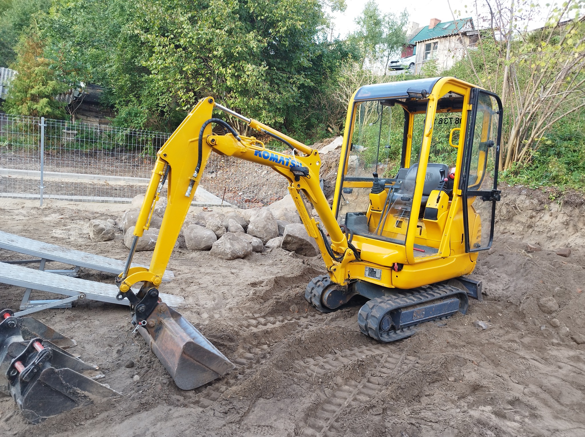 Żółta mini koparka Komatsu na placu budowy, obok leżą dodatkowe łyżki, a w tle widać kamienie i zieleń.