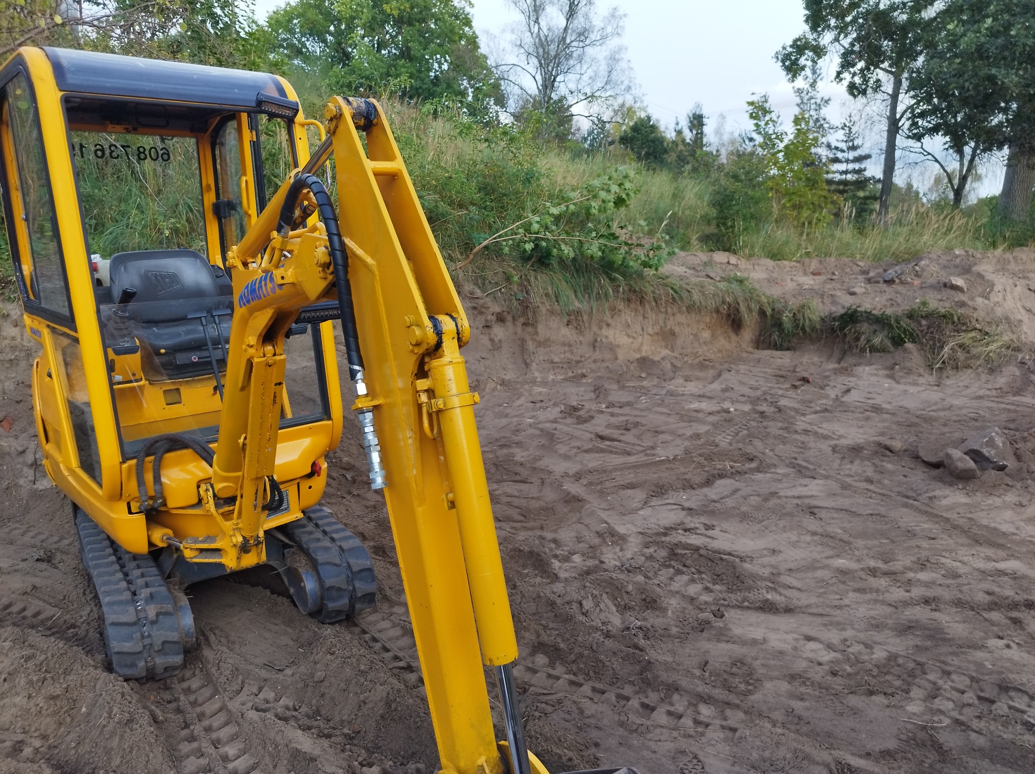 Żółta mini koparka na gąsienicach stoi na placu budowy z wykopanym zboczem ziemi w tle, widoczne ślady gąsienic na ziemi.