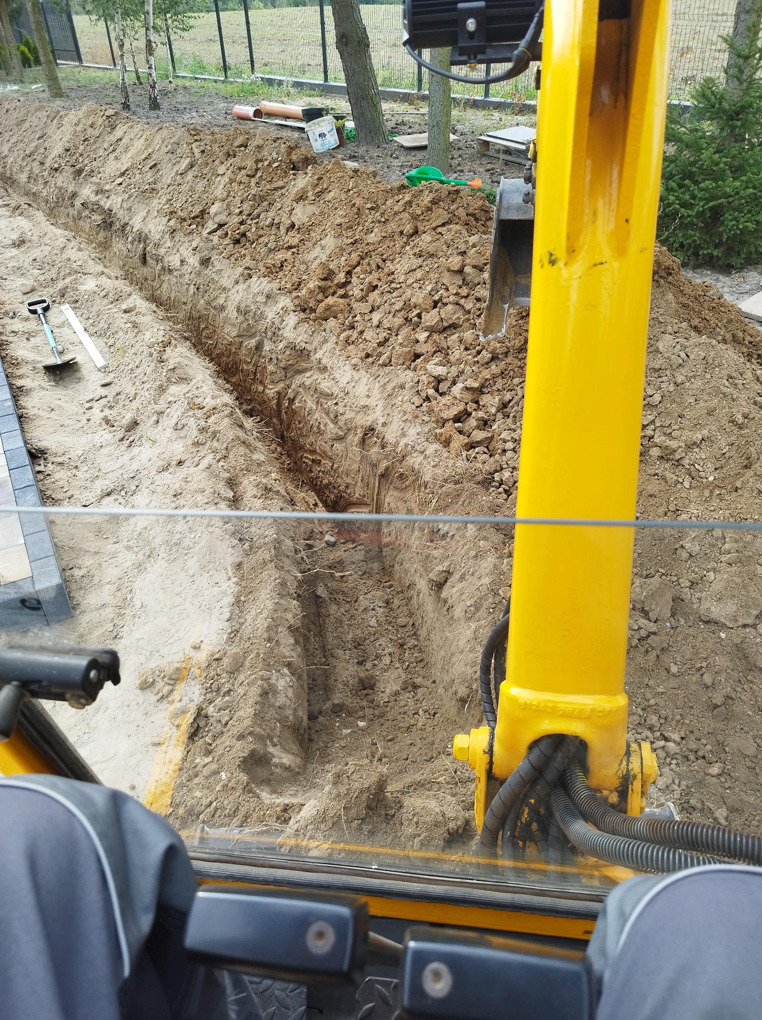 Wąski wykop pod instalacje widziany z kabiny żółtej koparki, w tle ułożona kostka brukowa i narzędzia.