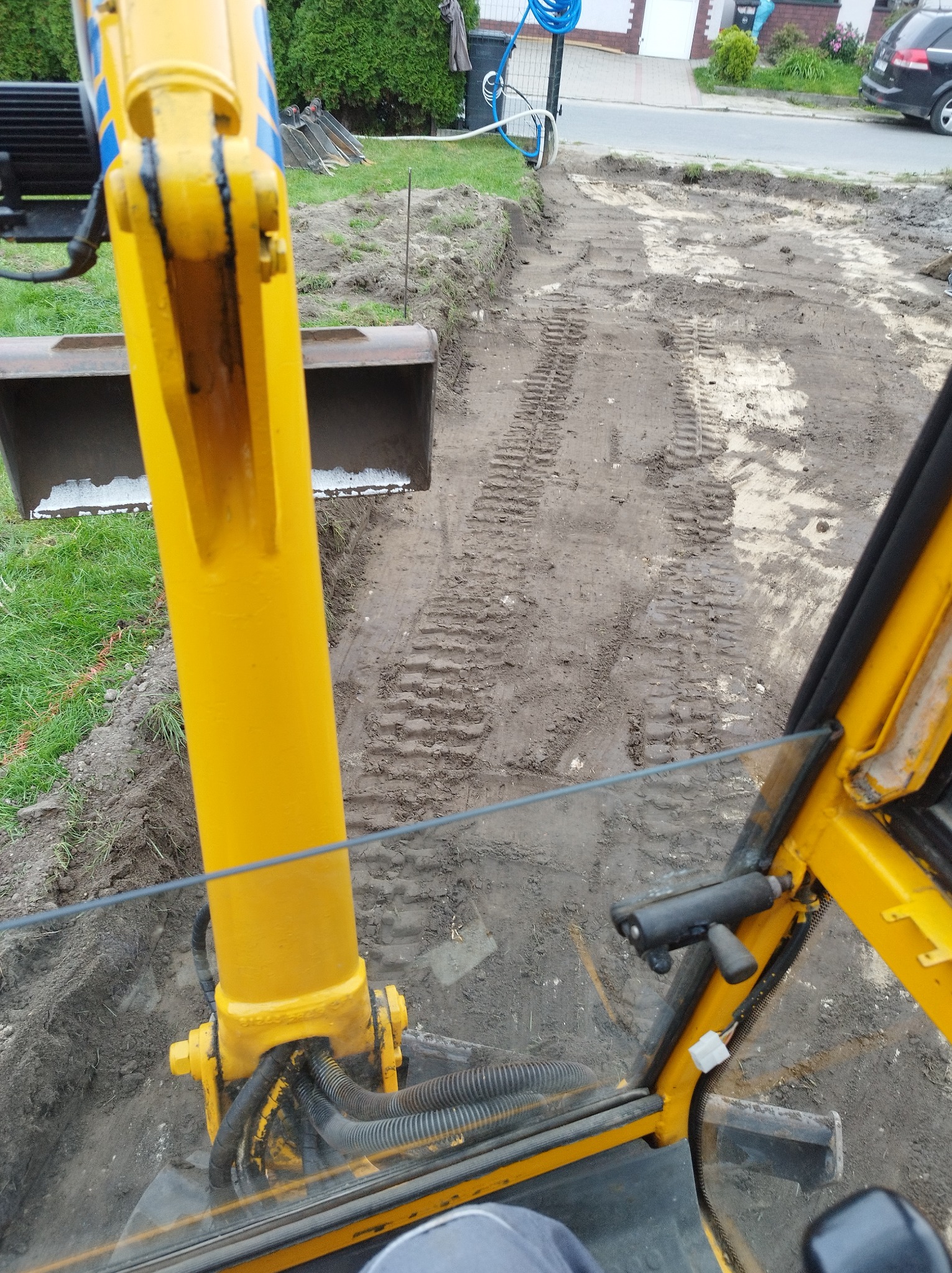 Widok z kabiny żółtej koparki na wykop w ziemi, widoczne ślady gąsienic i fragment zabudowy jednorodzinnej w tle.