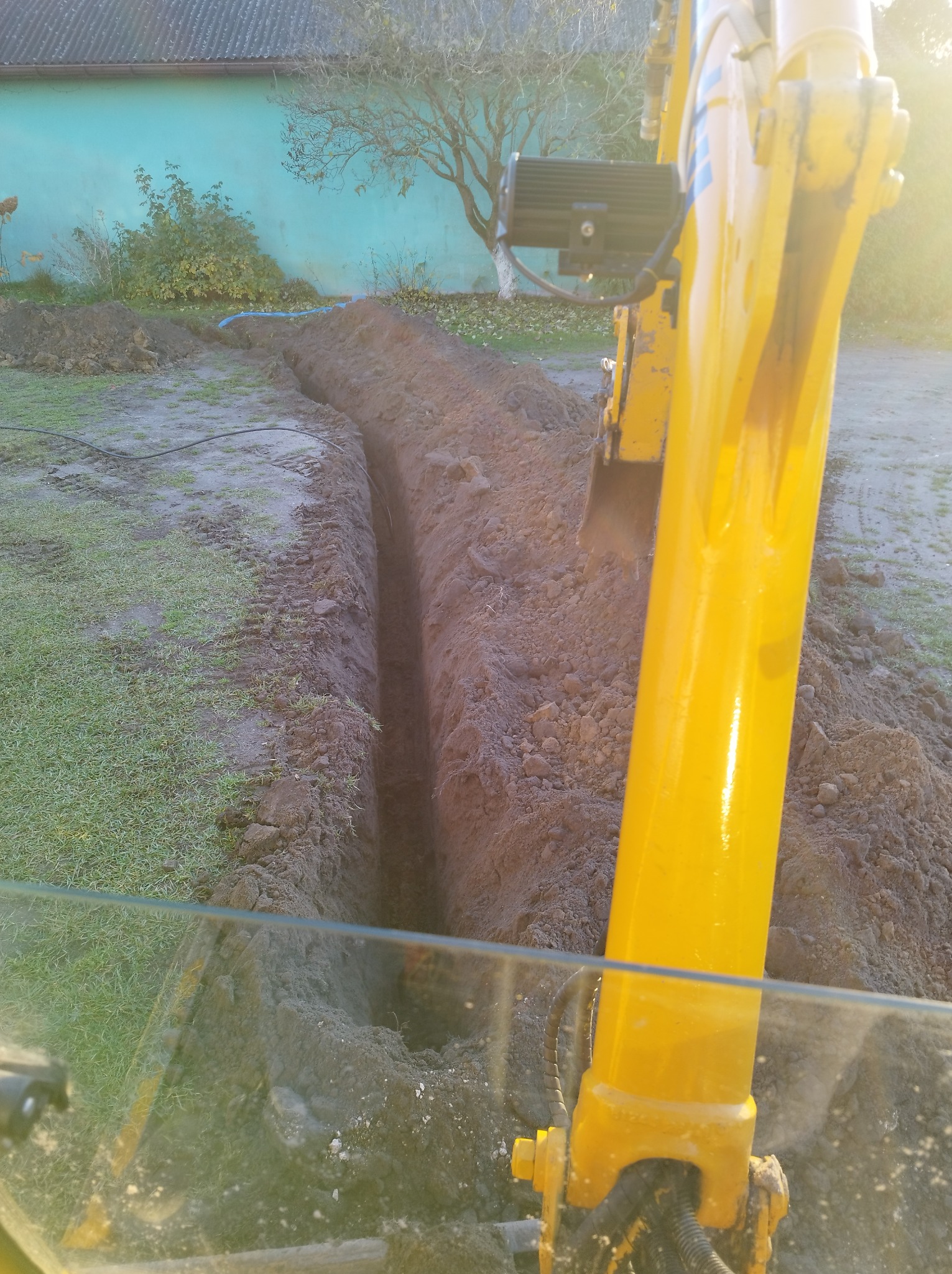 Żółta koparka w trakcie wykonywania wąskiego wykopu w ziemi, widoczne ułożone kable wzdłuż wykopu, w tle budynek z niebieską elewacją i drzewo.