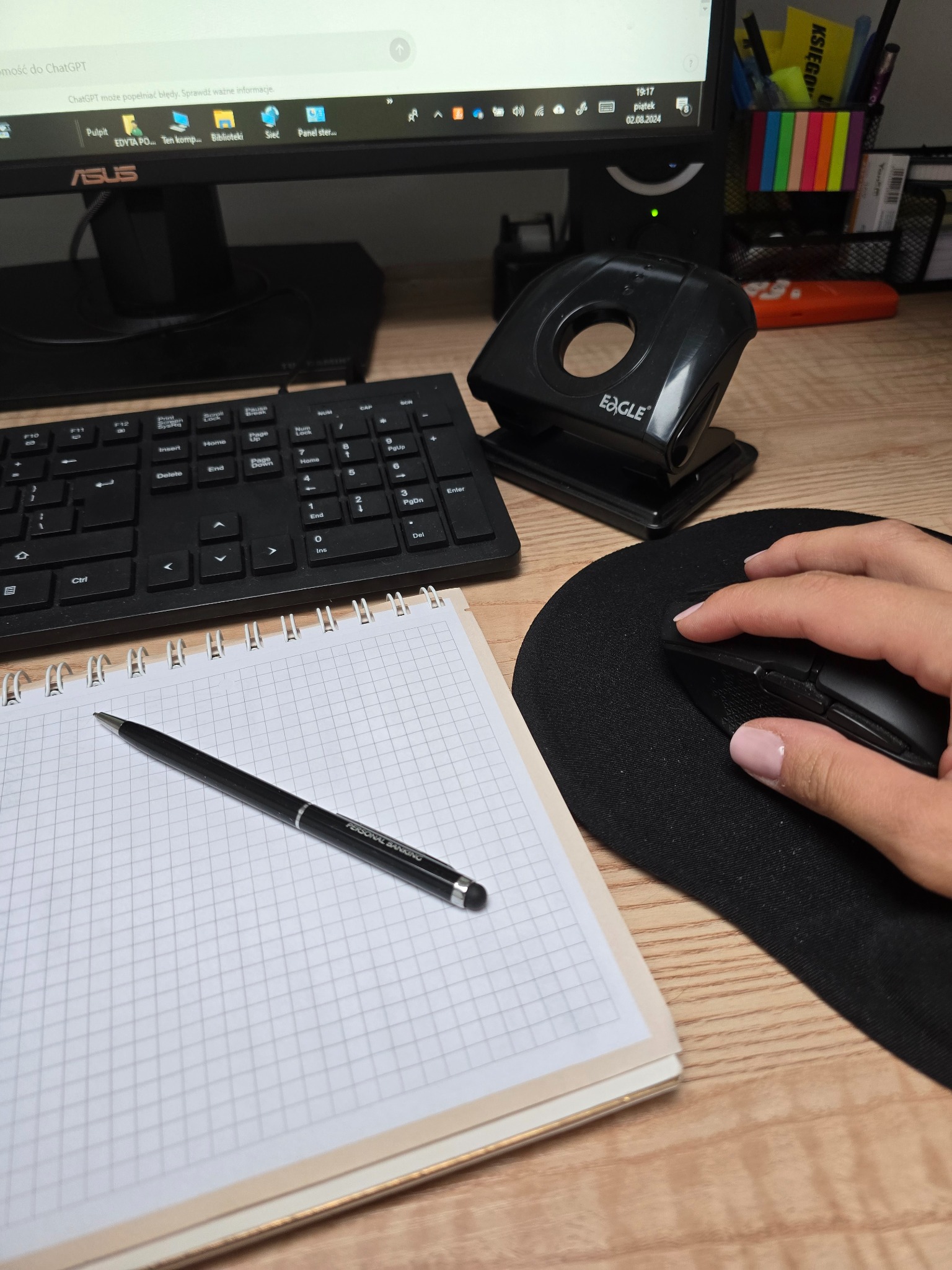 Biurko z komputerem, klawiaturą, dziurkaczem, podkładką pod mysz z dłonią, notatnikiem w kratkę i długopisem z napisem PERSONAL BANKING, sugerujące pracę biurową.