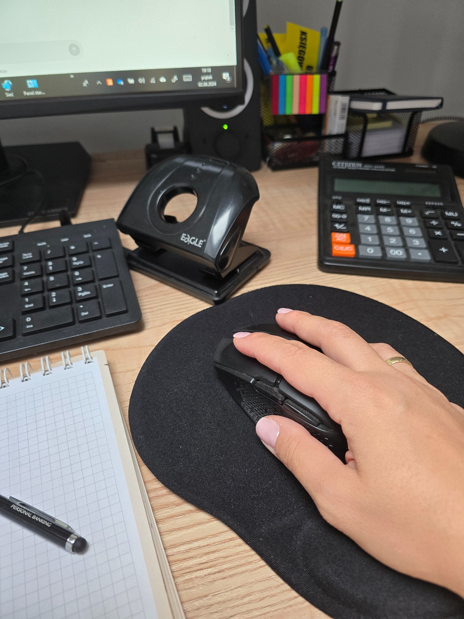 Dłoń kobiety z pierścionkiem na palcu serdecznym, obsługująca czarną myszkę komputerową na podkładce, biurko z kalkulatorem Citizen, dziurkaczem Eagle, klawiaturą numeryczną, notatnikiem...