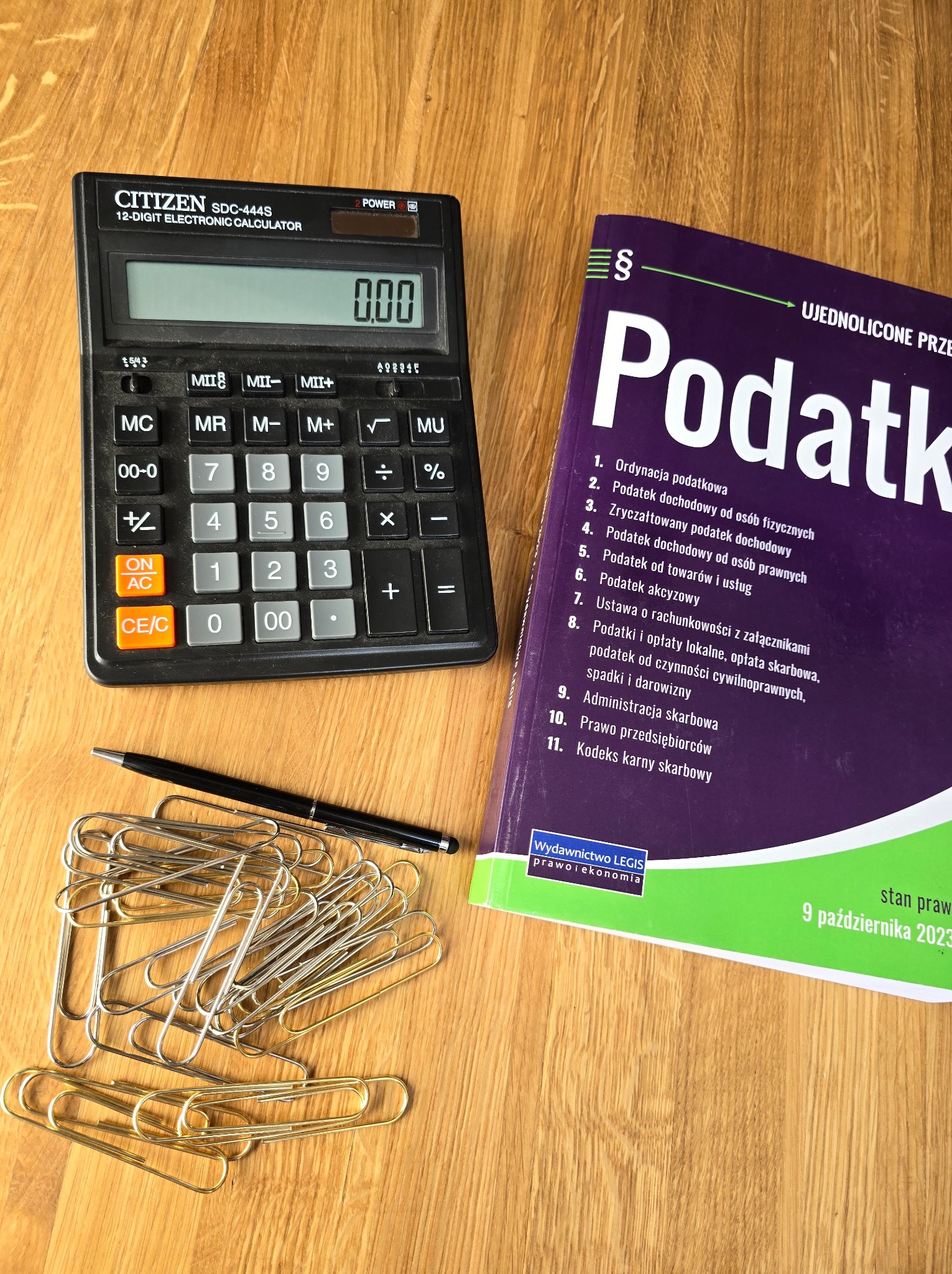 Kalkulator Citizen SDC-444S, sterta złotych spinaczy biurowych, czarny długopis i książka 'Podatki' Wydawnictwa Legis na drewnianym biurku.