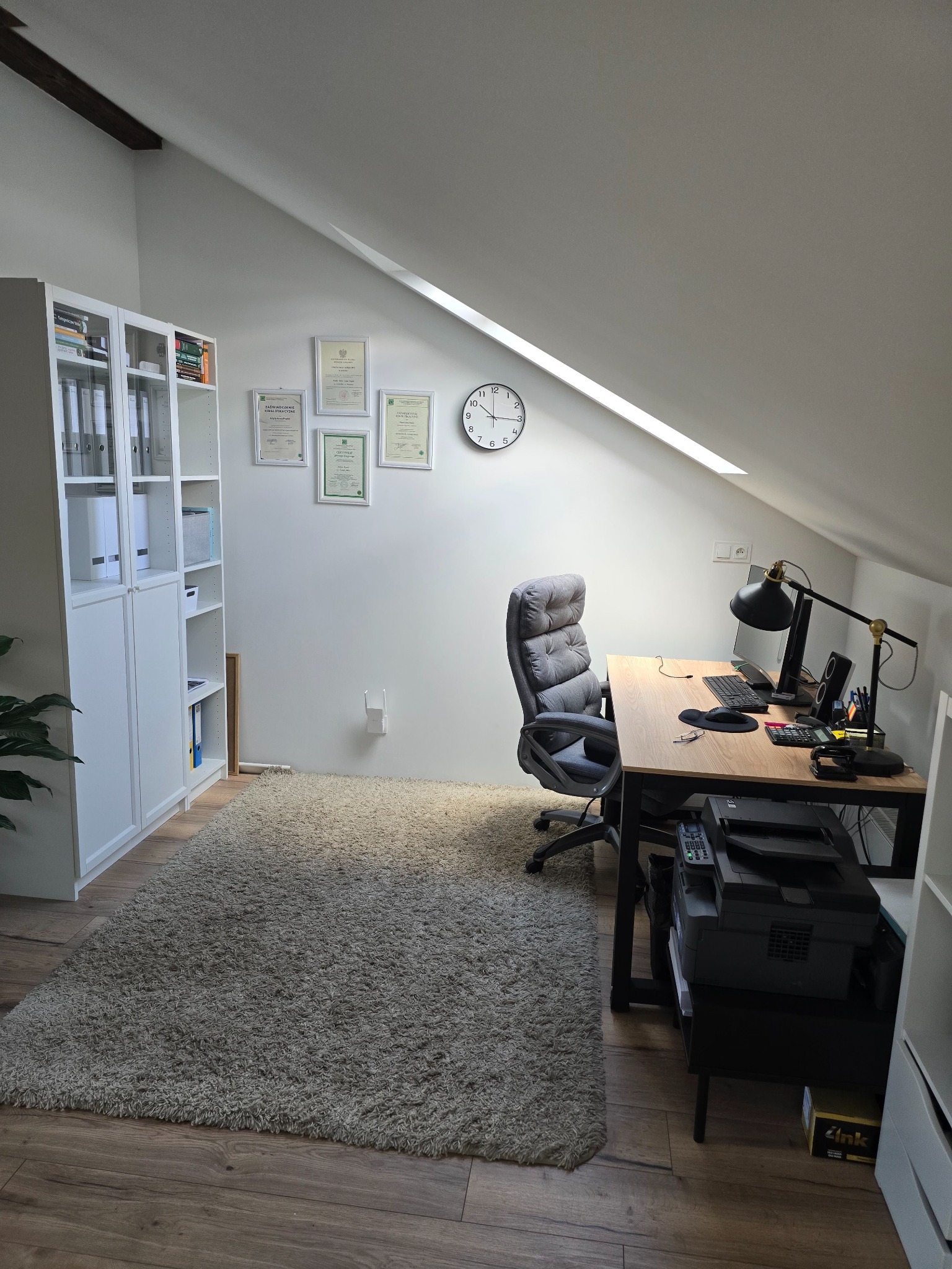 Biuro w pokoju na poddaszu z biurkiem, komputerem, krzesłem biurowym, szafą z segregatorami, dywanem i certyfikatami na ścianie.