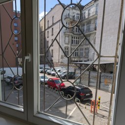 Kancelaria Radcy Prawnego Albert Białczyk - Widok z okna na parking z samochodami i budynek przez ozdobną kratę okienną. Słoneczny dzień, widoczne detale architektoniczne budynku.