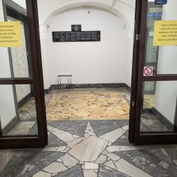 Kancelaria Radcy Prawnego Albert Białczyk - Wejście do budynku z tablicą informacyjną i posadzką w kształcie gwiazdy. Widok z zewnątrz przez otwarte, przeszklone drzwi.