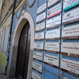 Kancelaria Radcy Prawnego Albert Białczyk - Tablica z szyldami różnych firm i usług, w tym kancelarii adwokackich, gabinetów psychologicznych i doradców finansowych, umieszczona na błękitnej ścianie budynku.